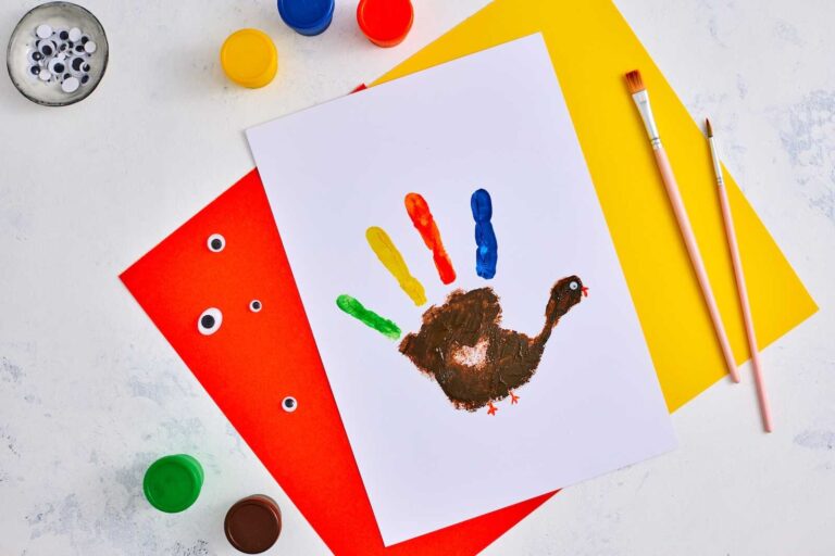 A colorful handprint turkey craft made with brown paint for the palm and thumb, and bright red, yellow, green, and blue paint for the feathered fingers, stamped onto white cardstock and surrounded by paint jars, googly eyes, and brushes on an orange and yellow background.