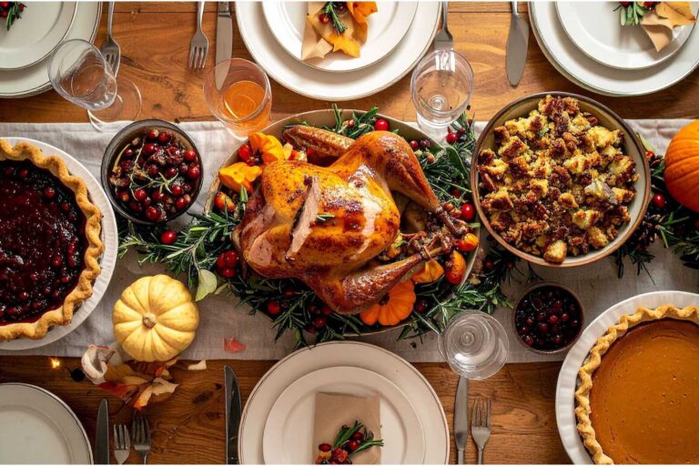 A Thanksgiving dinner table filled with a roasted turkey surrounded by fresh herbs and cranberries, bowls of stuffing and cranberry sauce, pumpkin pies, small decorative pumpkins, and neatly set place settings with plates, napkins, glasses, and silverware.