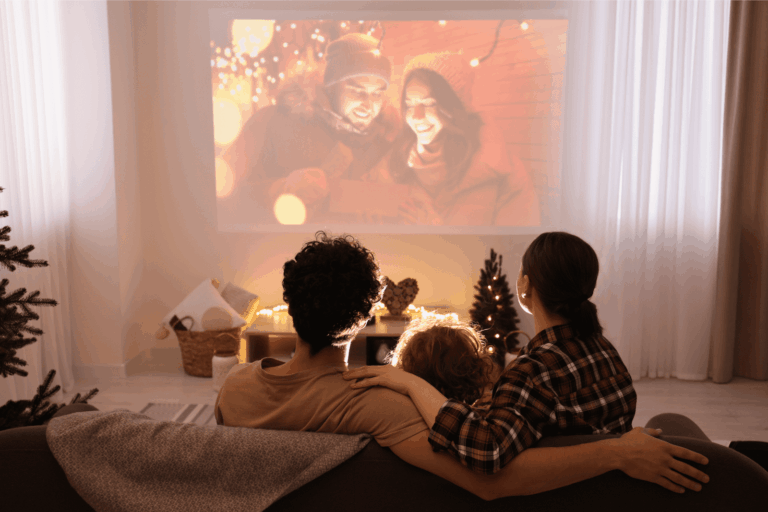 A family of three sits together on a couch in a softly lit living room, watching a holiday movie projected onto a large wall screen. The parents have their arms around their child as they look toward the warm, glowing scene on the screen, which shows a bundled-up couple surrounded by festive lights. Small Christmas trees, cozy blankets, and soft string lights decorate the room, creating a calm and inviting holiday atmosphere.