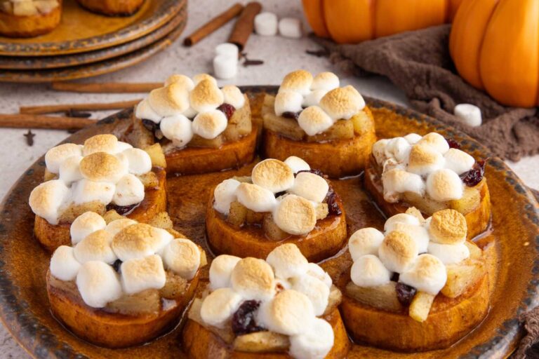 A rustic brown platter holds several thick, round slices of roasted sweet potato topped with golden-toasted mini marshmallows, diced pineapple, and dried cranberries, all glazed with a shiny cinnamon-honey sauce. In the background are cinnamon sticks, loose marshmallows, stacked plates, and small decorative pumpkins, creating a warm and cozy Thanksgiving atmosphere.