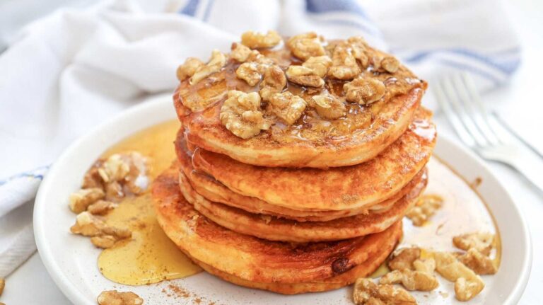 Stack of fluffy pumpkin pancakes topped with walnuts and maple syrup on a plate.