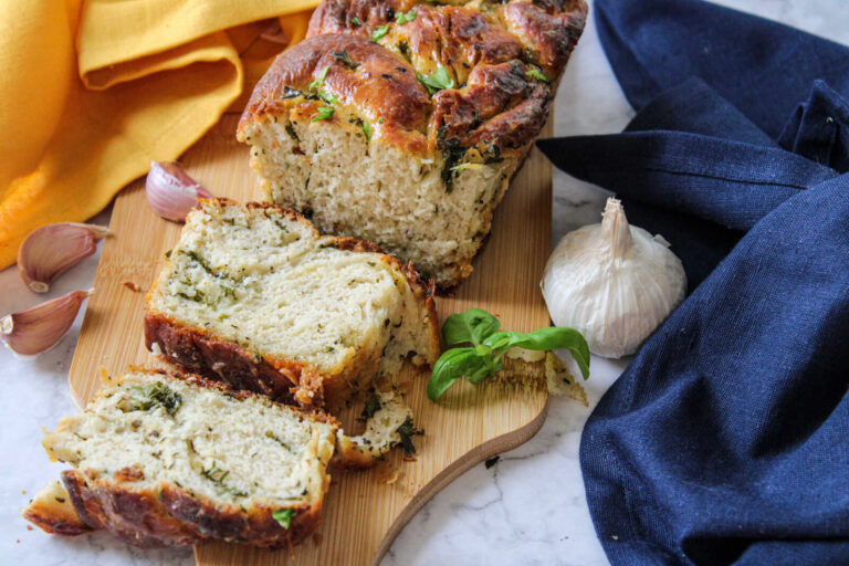 Sliced swirl garlic bread loaf with herbs on a wooden board alongside fresh garlic cloves.